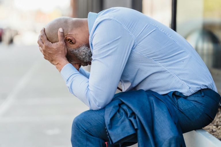 An adult man sitting outdoors, showing stress and contemplation with his head in hands.